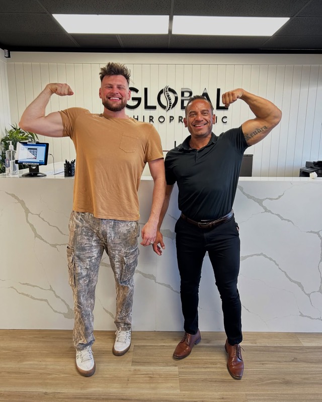 Dr. Steve and a patient flexing in front of the Global Chiropractic sign